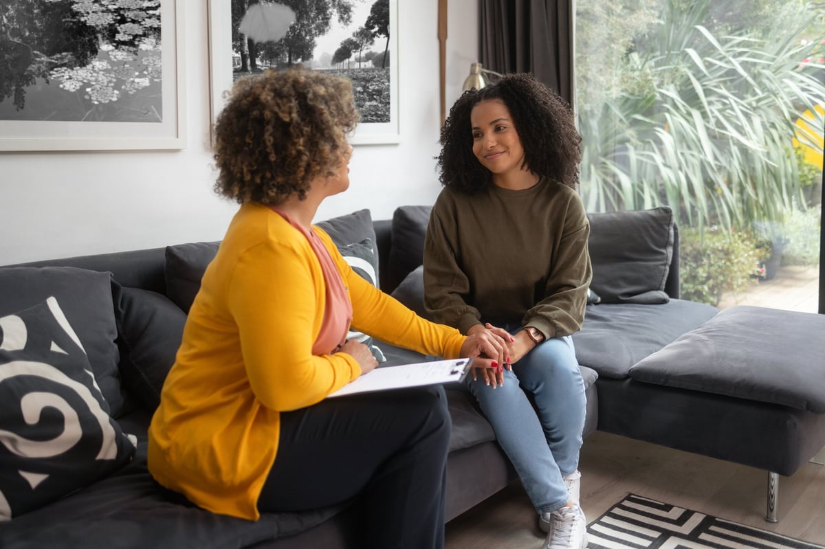 A female support worker sitting with a woman and calmly holding her hands during a supportive, confidential conversation, both faces fully visible in a warm and reassuring setting