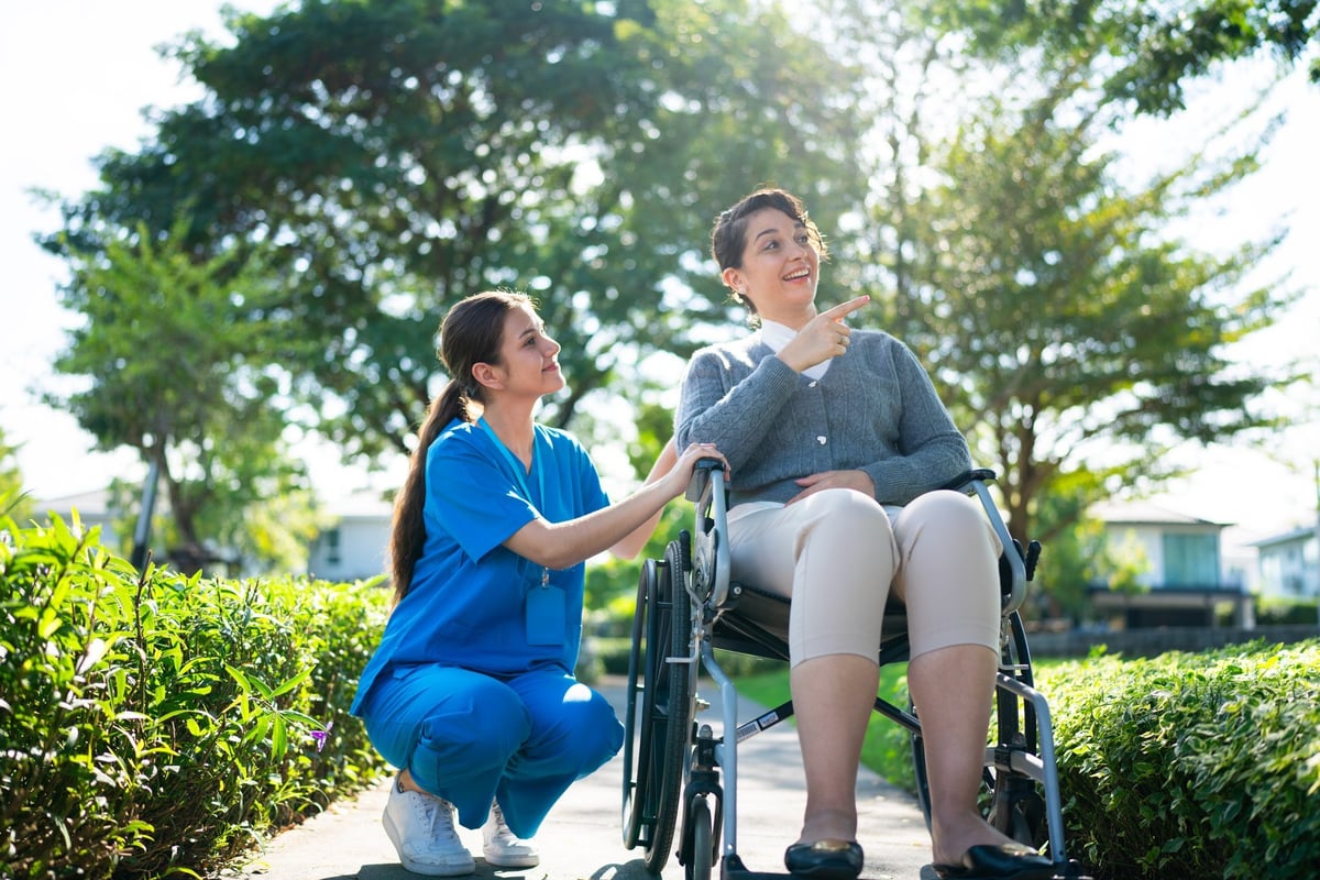 Nurse in blue scrubs kneeling beside a woman in a wheelchair, both with faces fully visible, engaging in conversation in a bright sunny park
