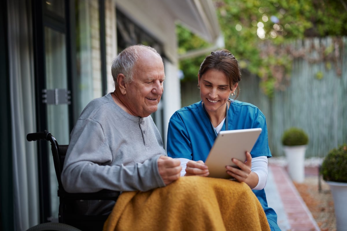 Careful nurse assisting a senior man while using a digital tablet on a patio, both smiling with faces fully visible while reviewing information together