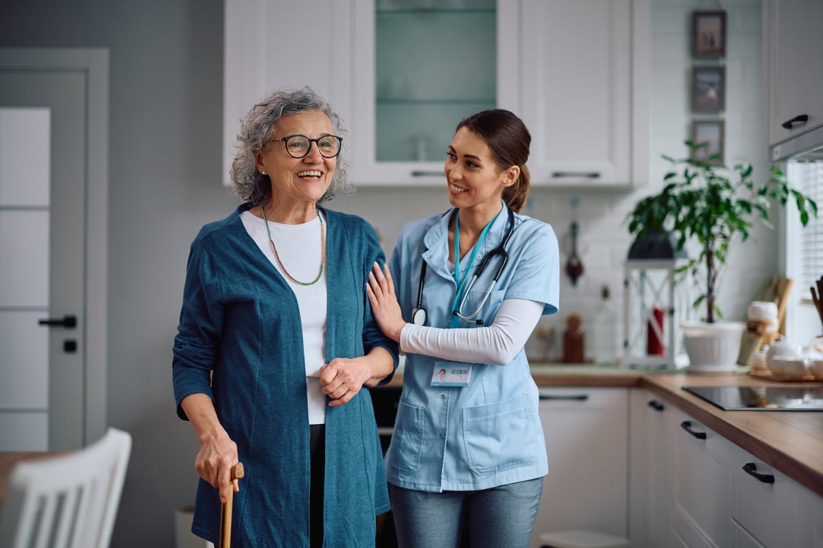 Happy home caregiver assisting her senior patient with walking through the kitchen, both smiling with faces fully visible in a bright home setting