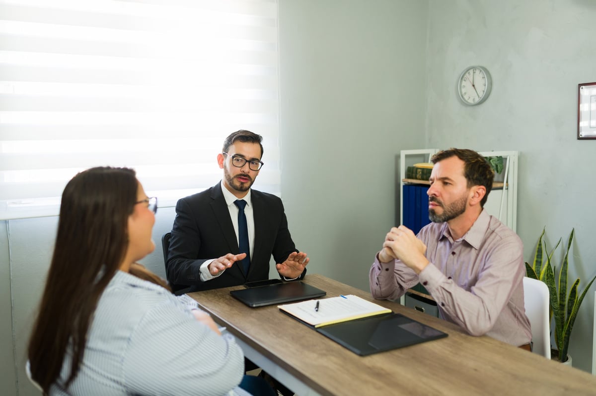 Couple in a constructive facilitated mediation meeting with a practitioner explaining options, all faces fully visible, in a calm office setting