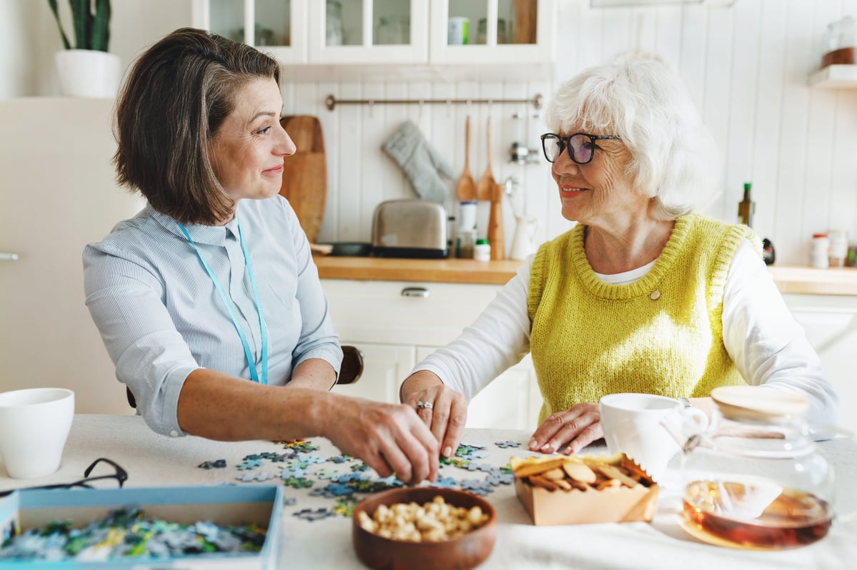 Caregiver sitting with an older woman in glasses completing a puzzle together at a kitchen table, supporting dementia cognitive care in a warm home setting