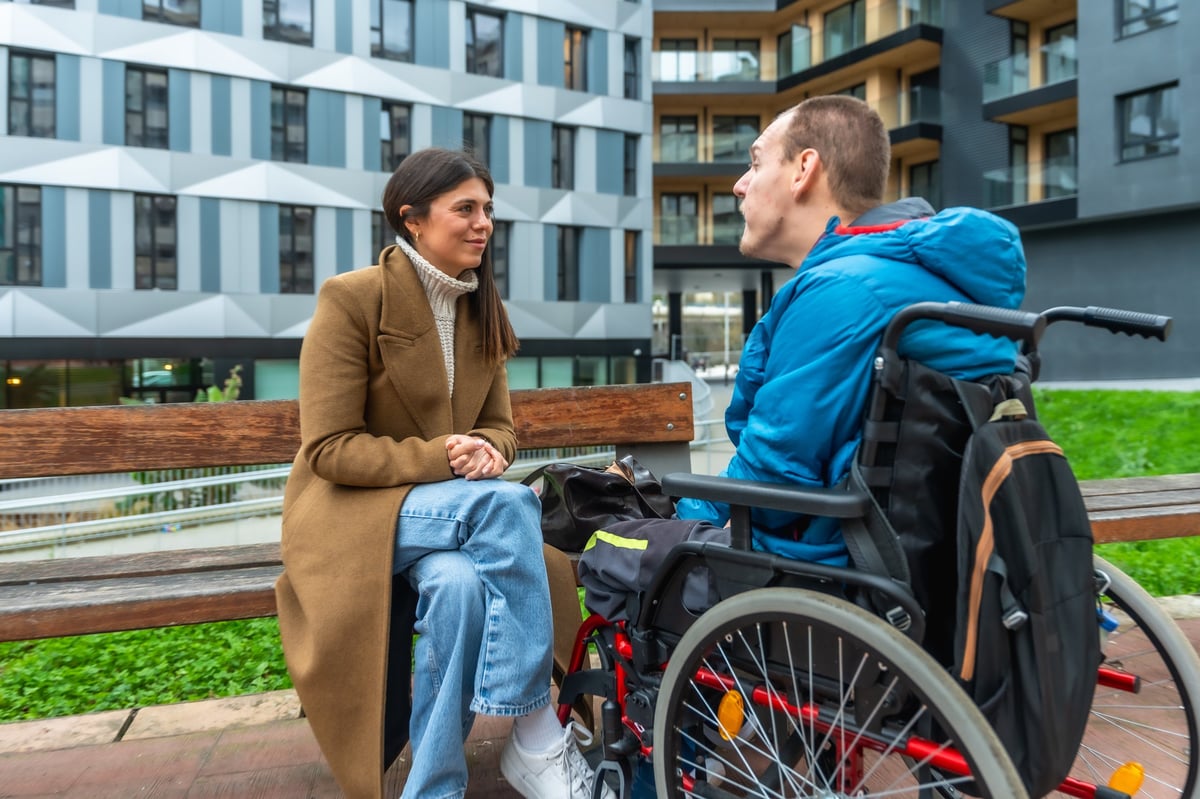 Support practitioner talking with a person using a wheelchair on a park bench, both with heads and faces fully visible