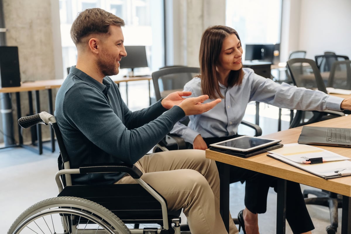 Man in a wheelchair talks with a woman in a blue shirt at a desk in a contemporary office, featuring a laptop and tablet on the surface