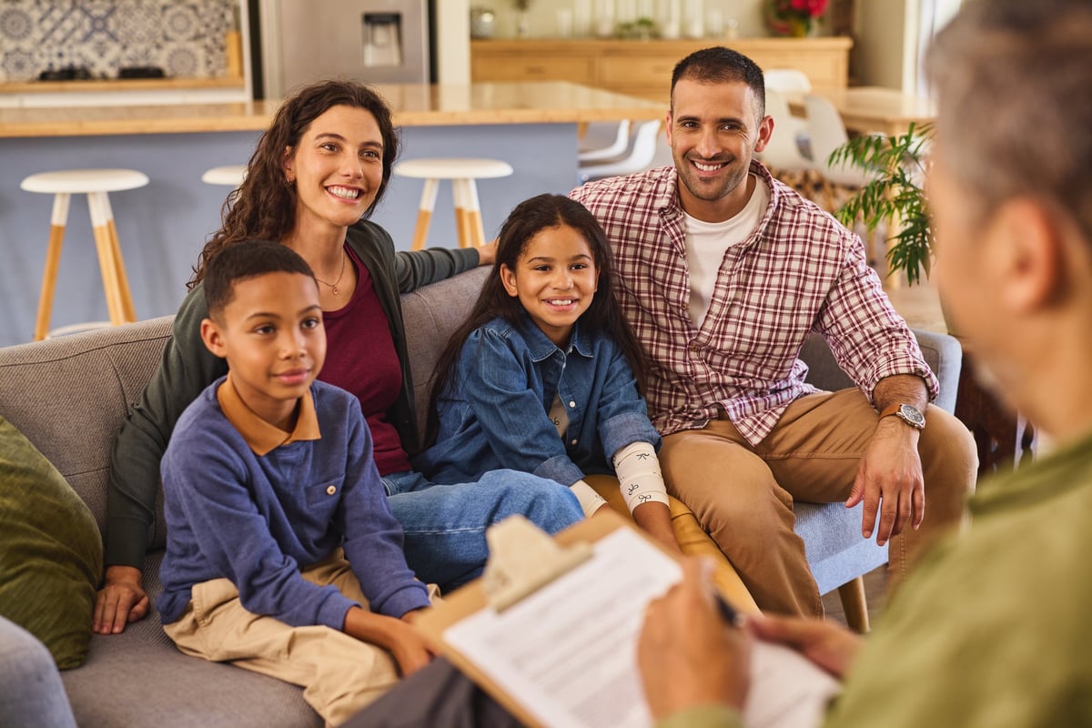 Multicultural family sitting on a couch talking with a friendly support worker, all faces visible, in a warm home consultation setting