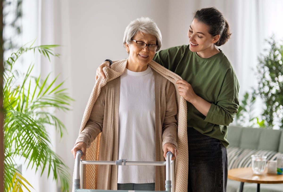 Cheerful caregiver gently placing a cozy shawl on an elderly woman with a walker in a bright living room, capturing warmth, trust and in-home aged care support