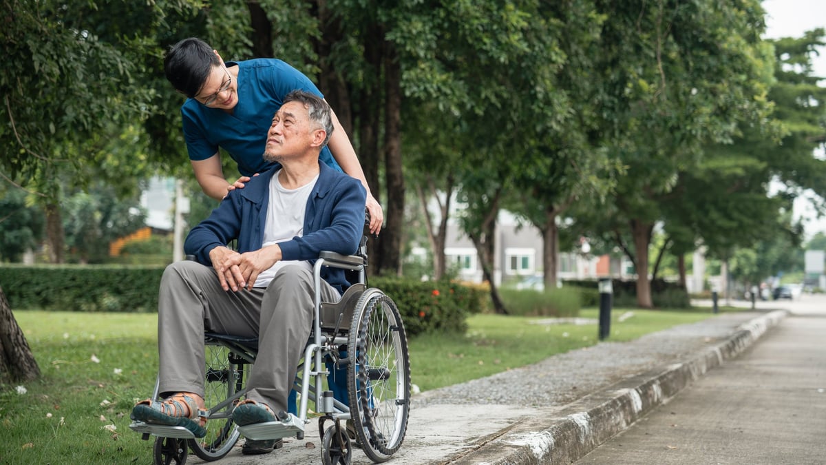 Support worker guiding a participant in a wheelchair along a paved park path, both with heads and faces fully visible